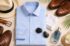 Flat lay of a light blue men’s dress shirt with sunglasses, hat, loafers, belt, watch, cologne and tropical leaves styled for hot, humid weather.