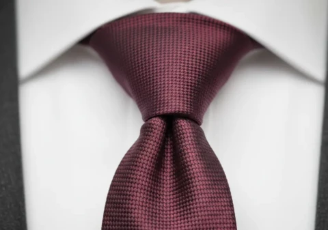 Close-up of a Windsor knot in a burgundy grenadine tie against a white dress shirt collar with a charcoal suit lapel visible