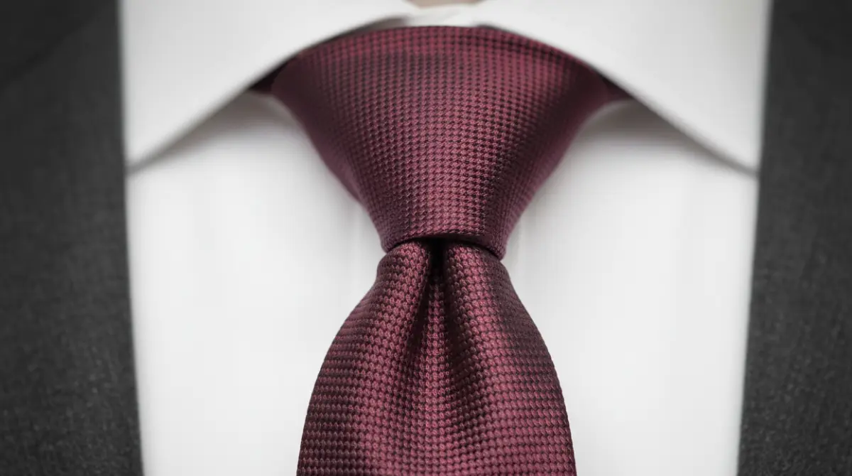 Close-up of a Windsor knot in a burgundy grenadine tie against a white dress shirt collar with a charcoal suit lapel visible