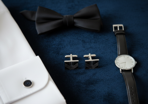 Flat lay of silver cufflinks, white French cuff dress shirt, black bow tie, and silver watch on navy velvet surface